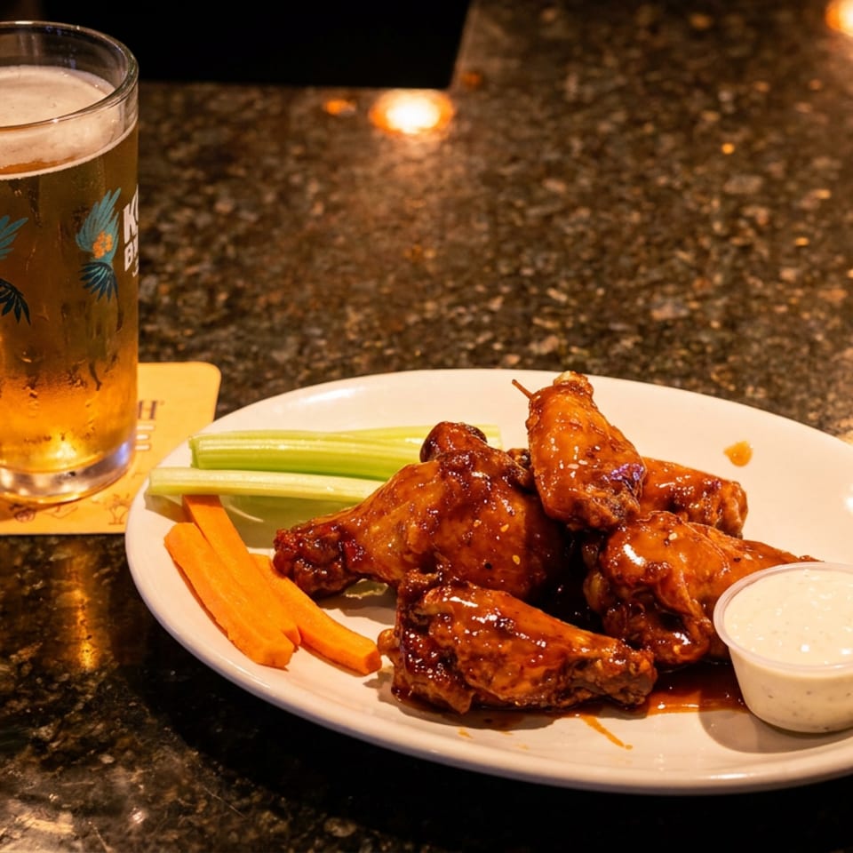 Plate of saucy chicken wings with celery, carrots, and dipping sauce beside a pint of beer on a dark tabletop.