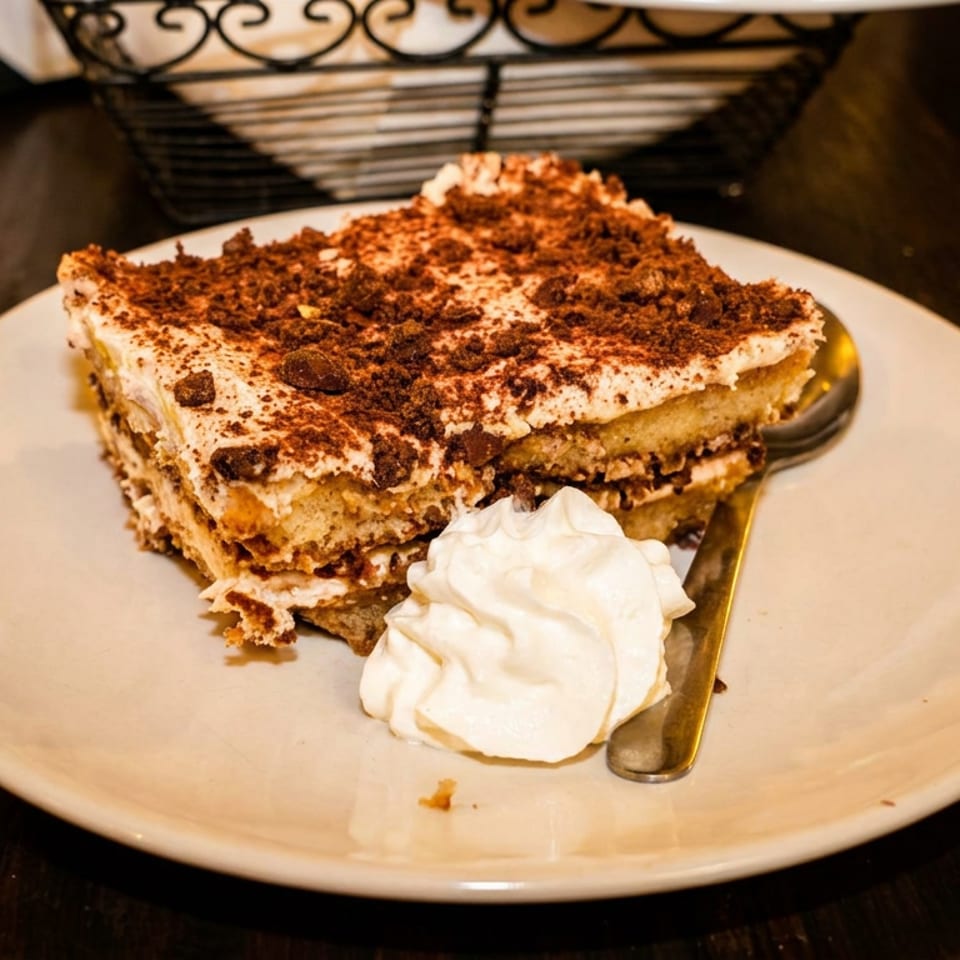 Slice of cocoa-dusted tiramisu with whipped cream and spoon on a white plate at a warmly lit restaurant table.